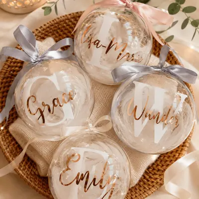 Cuatro bolas de Navidad transparentes con plumas dentro, la inicial en blanco y el nombre en dorado rosado