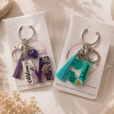 Dos llaveros de letra en resina con flores naturales deshidratadas, glitter y borla de hilo, en su empaque de marca