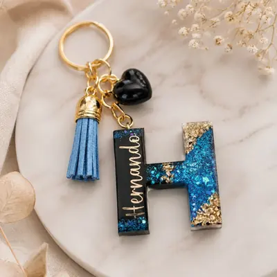Llavero de resina con la letra H en negro, glitter azul y hoja de oro, nombre en dorado, corazón negro y borla azul